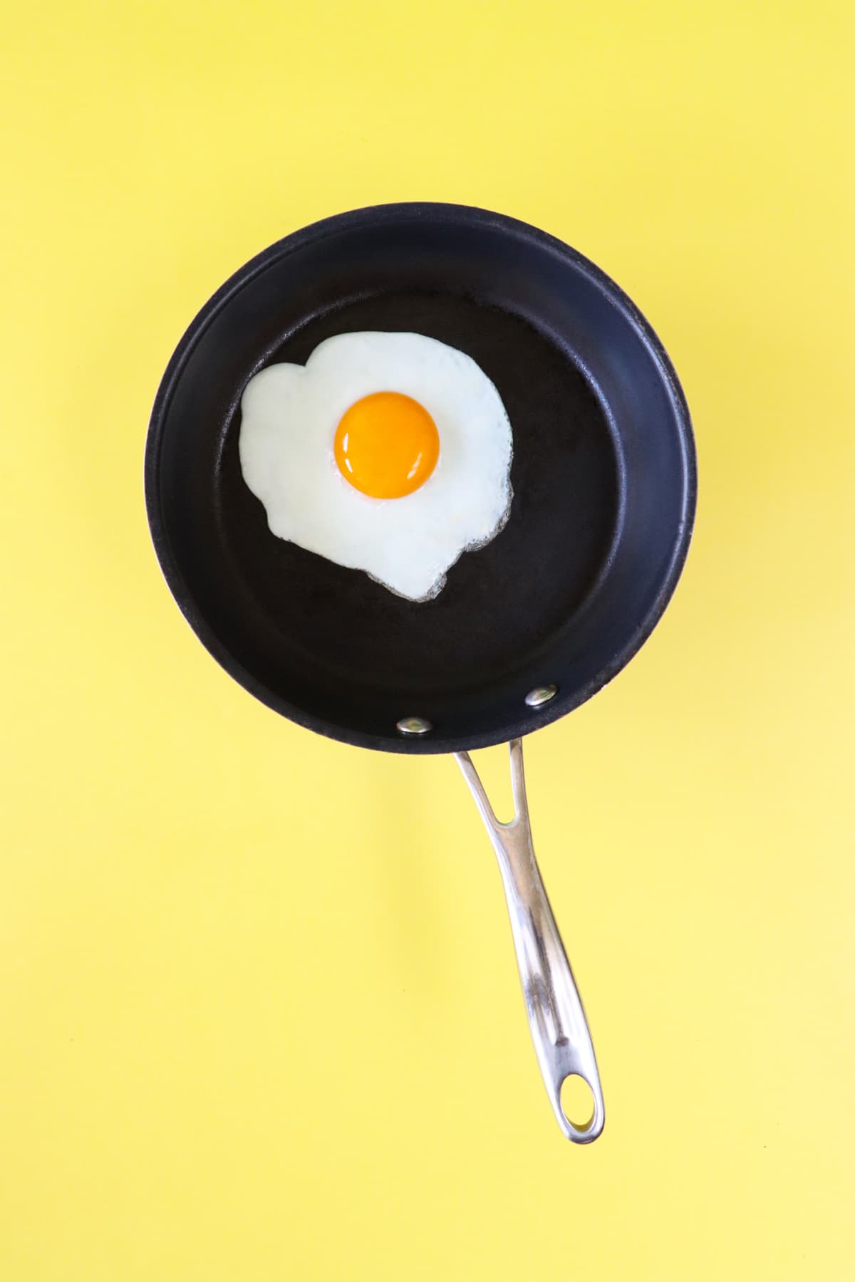 Stock photo showing an elevated view of sunny side up fried egg in a non-stick frying pan against a yellow background.