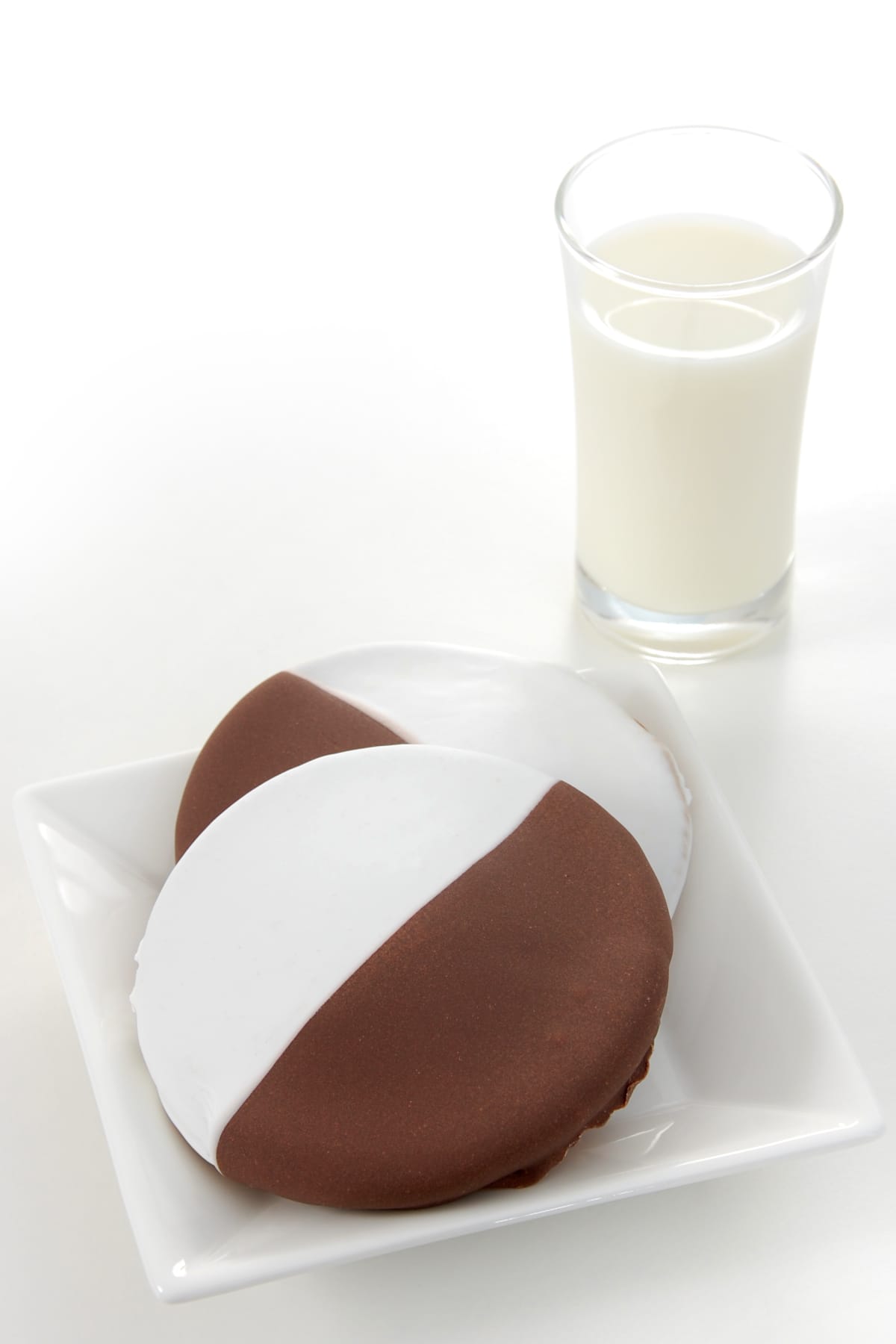 Two "half-moons" or "black and white" cookies frosted with chocolate and vanilla fondant, served with a glass of milk.