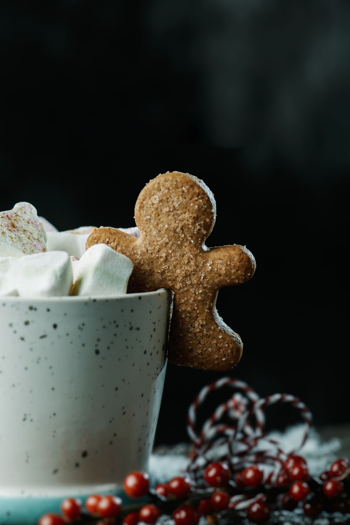 closeup of a gingerbread man cookie in a cup of coffee topped with some marshmallows with different shapes, on a gray rustic wooden table, against a dark gray backgrounw with some blank space on top