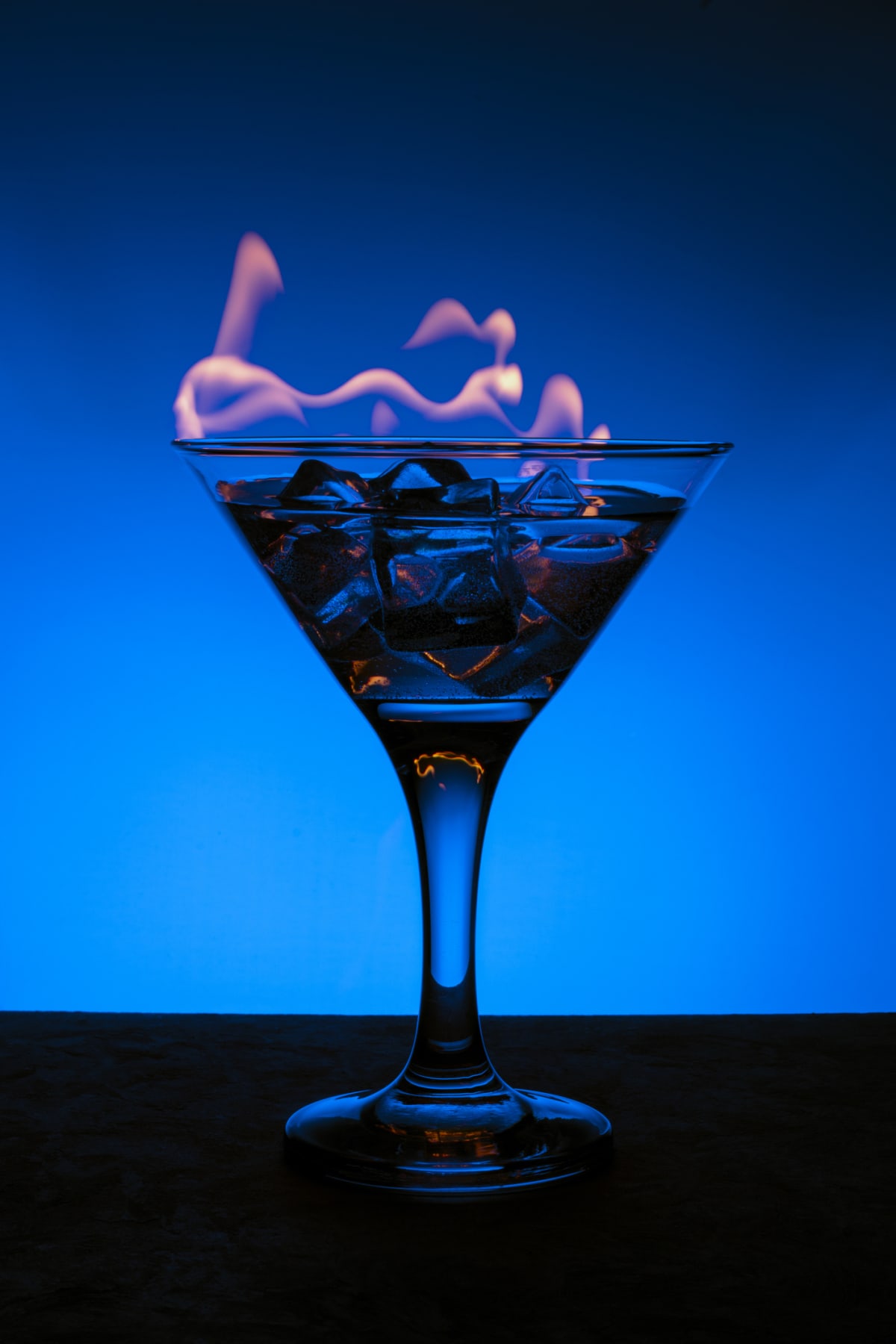 Burning alcoholic drink with ice cubes, on a blue gradient background, vertical frame. Burning cocktail on table in a bar. Blue alcoholic beverage and fire.