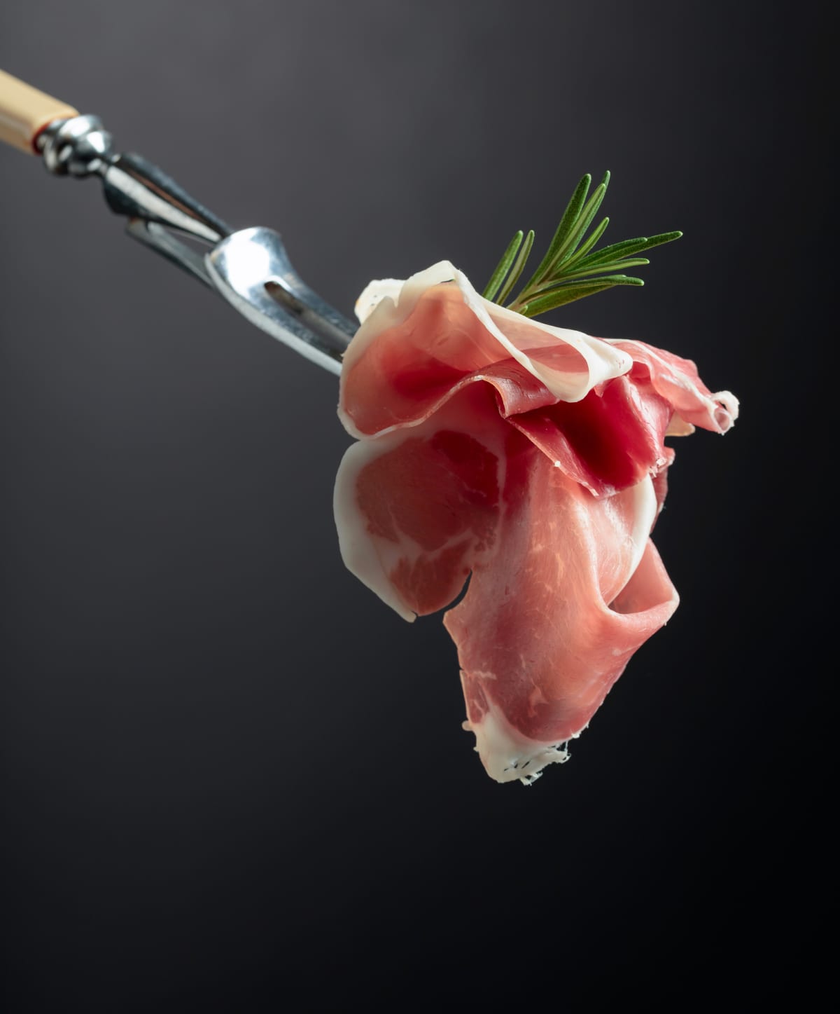 Sliced prosciutto with rosemary on a fork, black background, copy space.