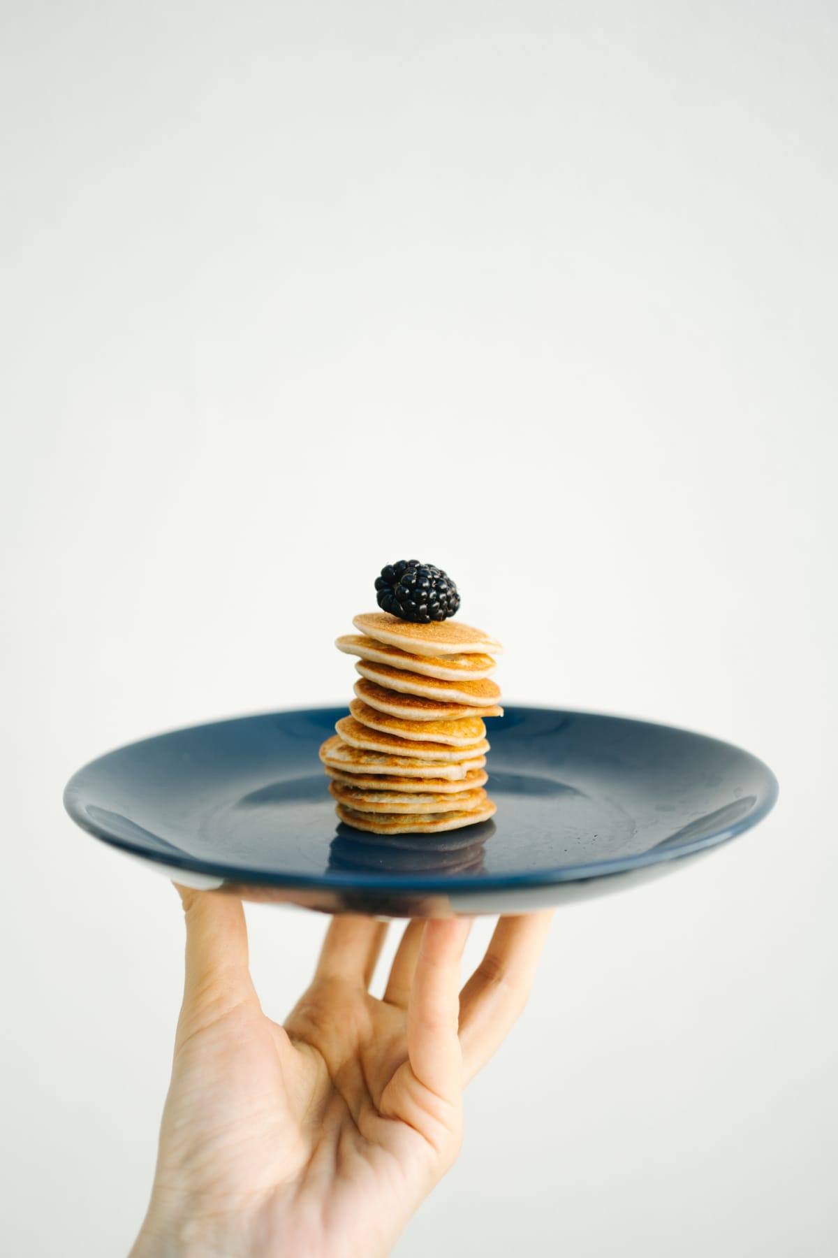 a hand holing a blue plate that has a stack of mini pancakes with a blackberry on top