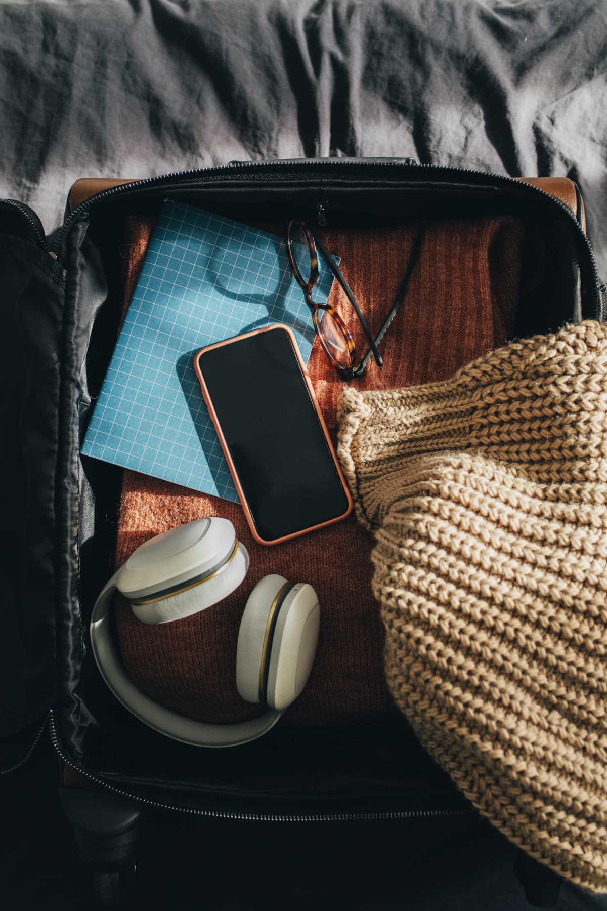 Woman's suitcase laid on a bed with a lot of travel-related objects all around: a mobile phone, clothes, accessories.