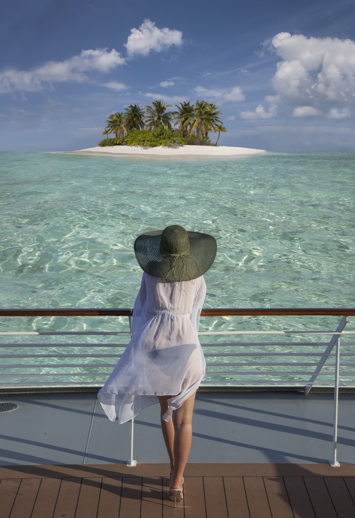 A young woman on the deck of a cruising ship watching a small island with white sands and coconut palms.