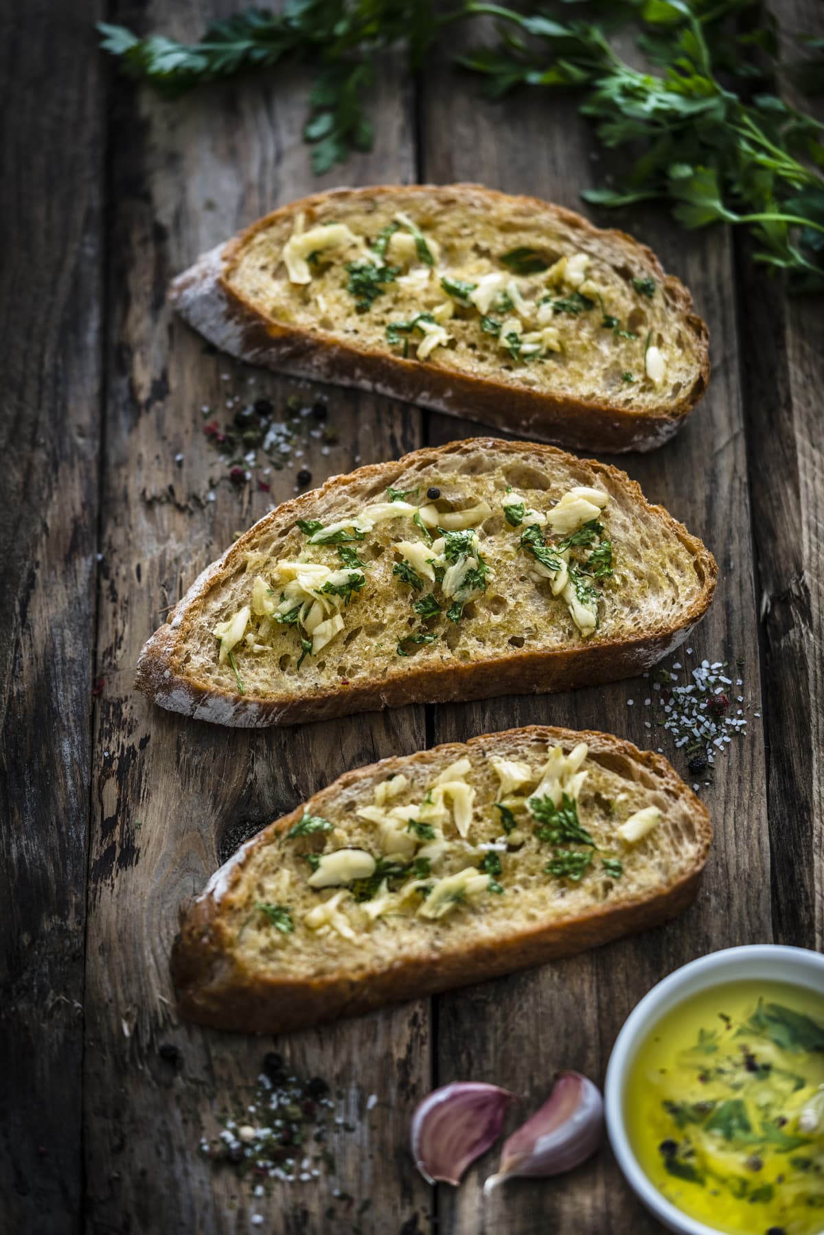 Appetizer: garlic bread on rustic wooden table. Three toasted bread slices with garlic and parsley on rustic wooden table. Parsley bunch, garlic cloves and olive oil complete the composition. Predominant colors are yellow and brown. XXXL 42Mp studio photo taken with SONY A7rII and Zeiss Batis 40mm F2.0 CF