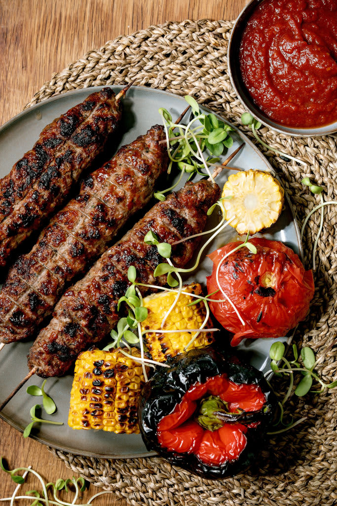 Eastern fast food. Grilled spicy beef lyulya kebab on sticks on plate with grilled vegetables sweet corn cob, tomato and paprika, tomato sauce on wooden table. Flat lay. (Photo by: Natasha Breen/REDA&CO/Universal Images Group via Getty Images)