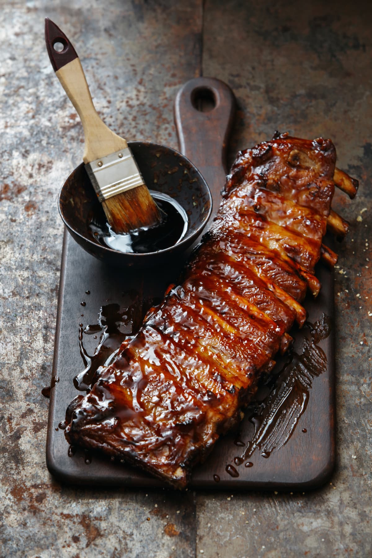 Pork ribs on a cutting board next to a bowl of sauce and a marinade brush