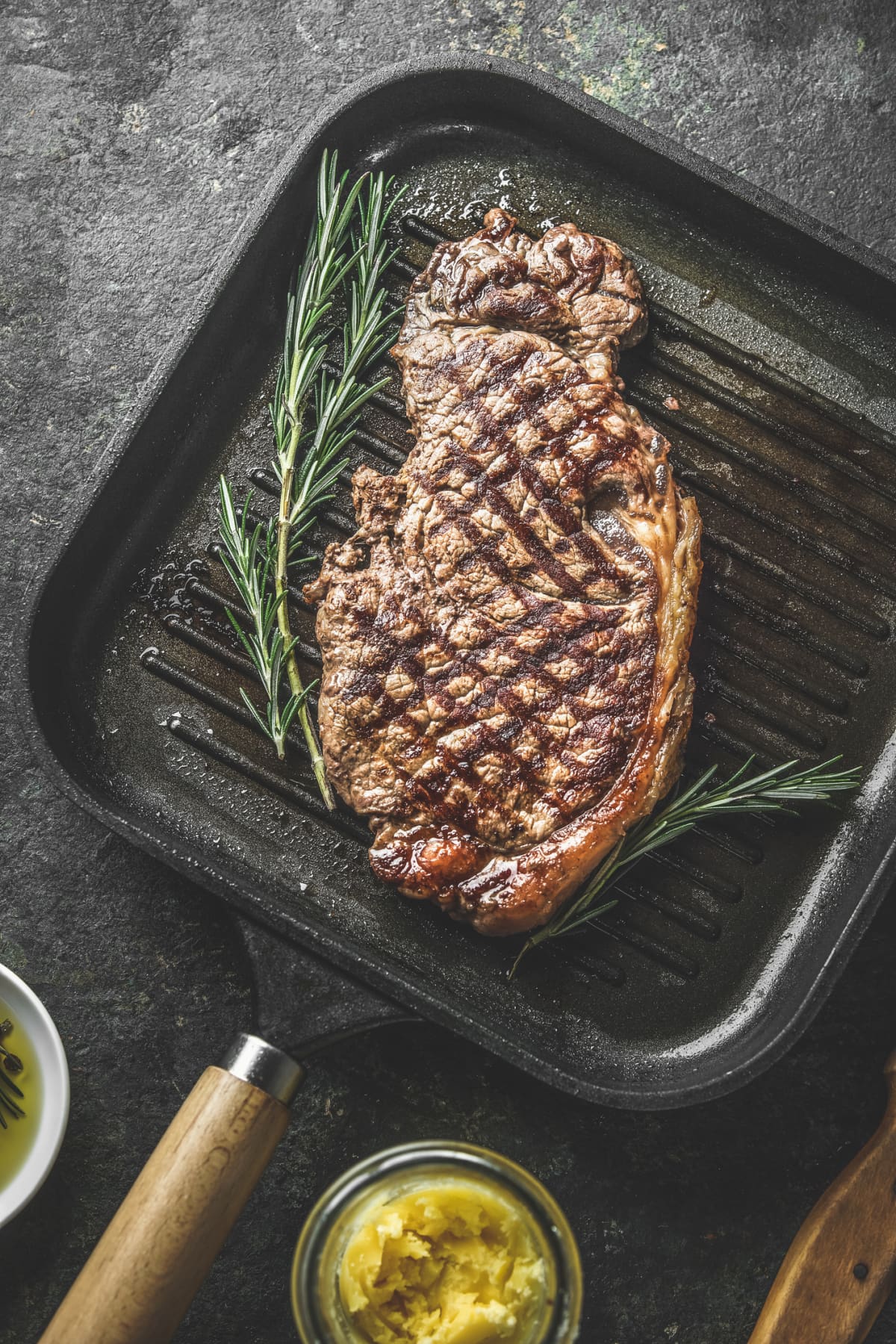 A large cooked ribeye steak in a grill pan next to a sprig of rosemary