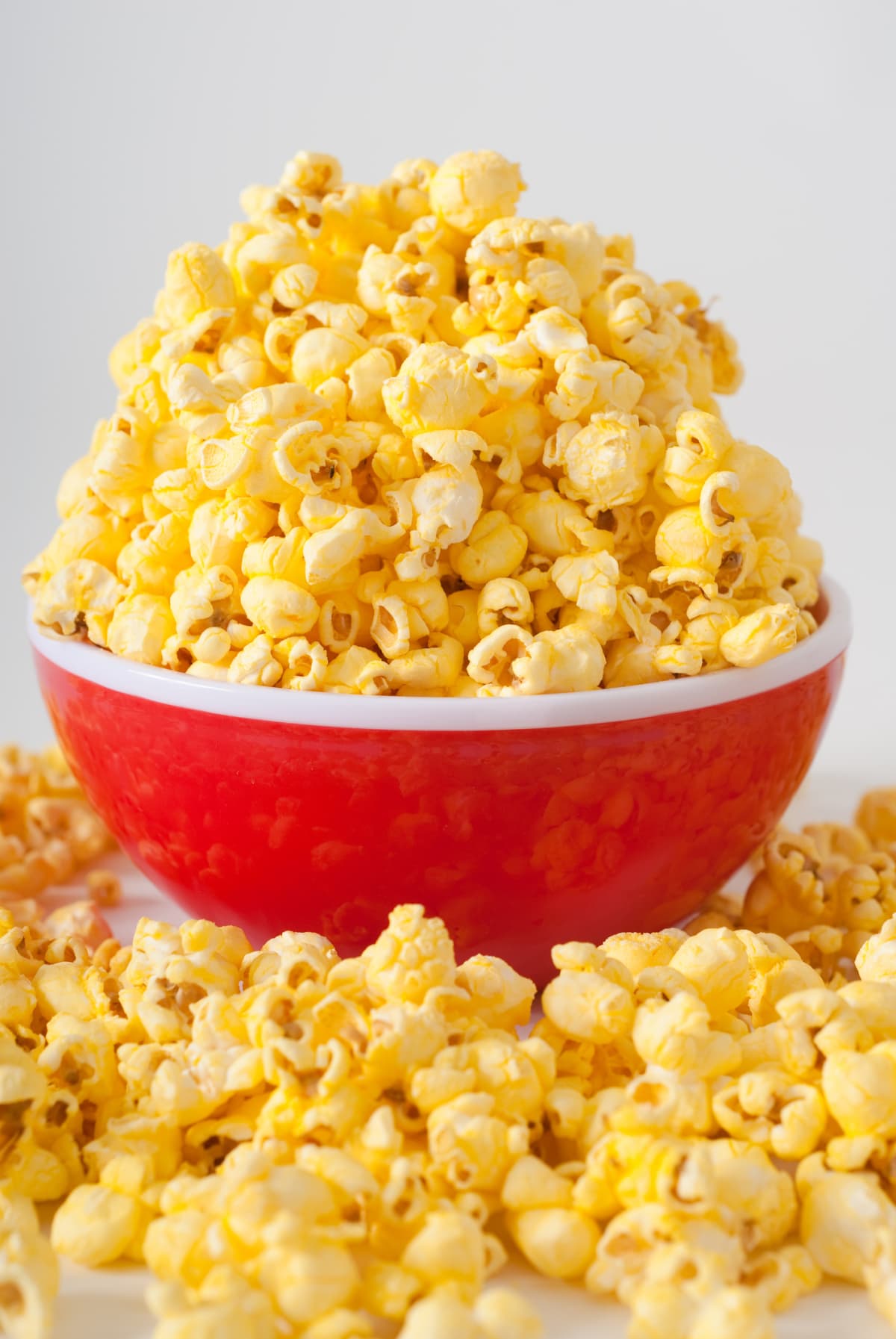 Popcorn piled high in a white and red bowl on a white surface with popcorn all around