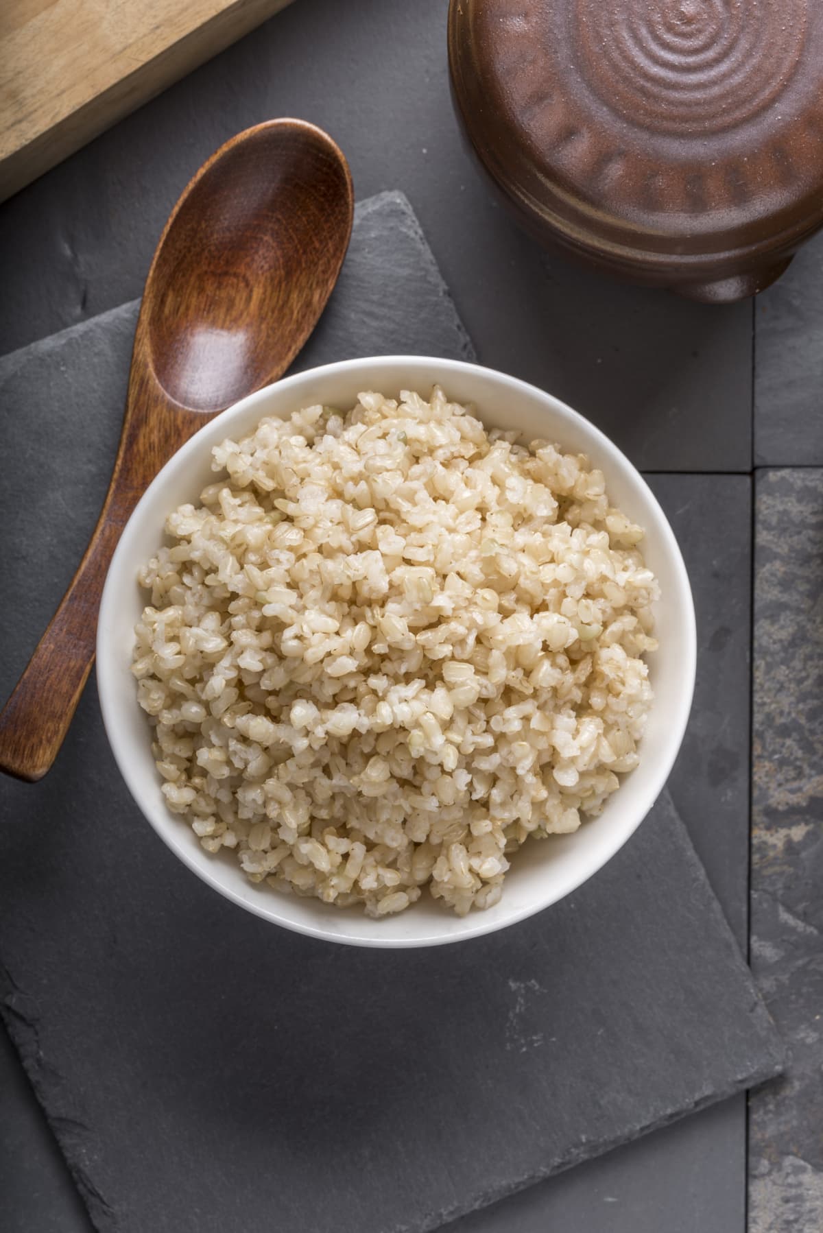 Cooked brown rice in a white bowl next to a wooden spoon on black surface