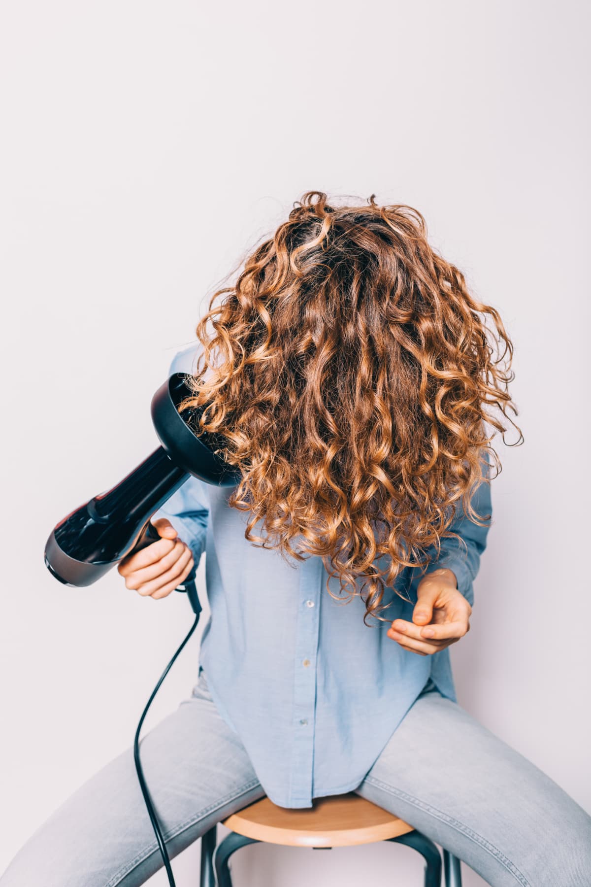 Young woman sitting on chair styling her curly hair with hairdryer with special diffuser nozzle.