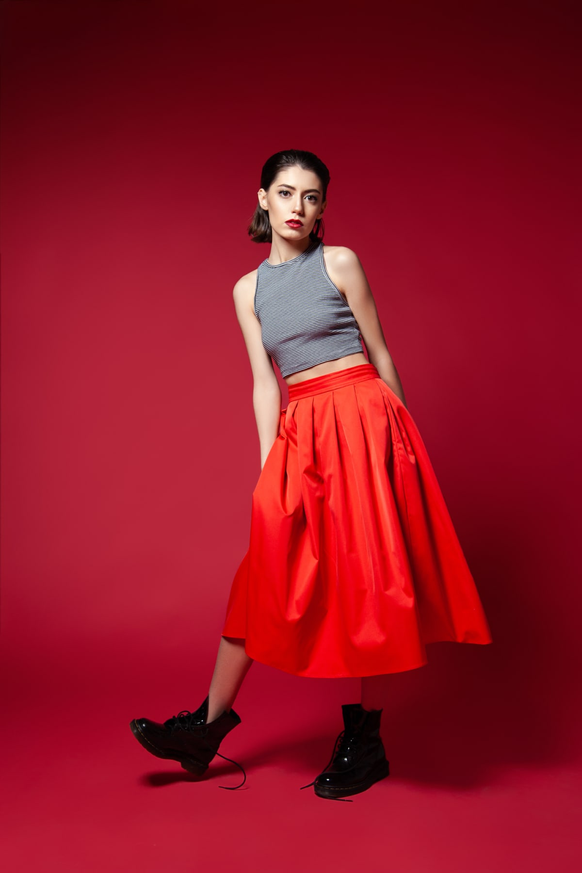 Young slender brunette in long red skirt and black boots balancing on one leg looking at camera on red background