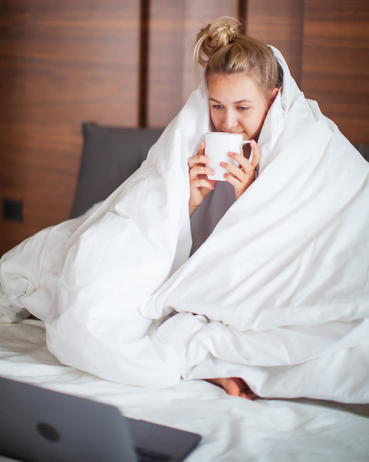 Cute smiling teen girl under weighted blanket in white bed in bedroom woken up and drink tea. Breakfast in bed, lazy morning.