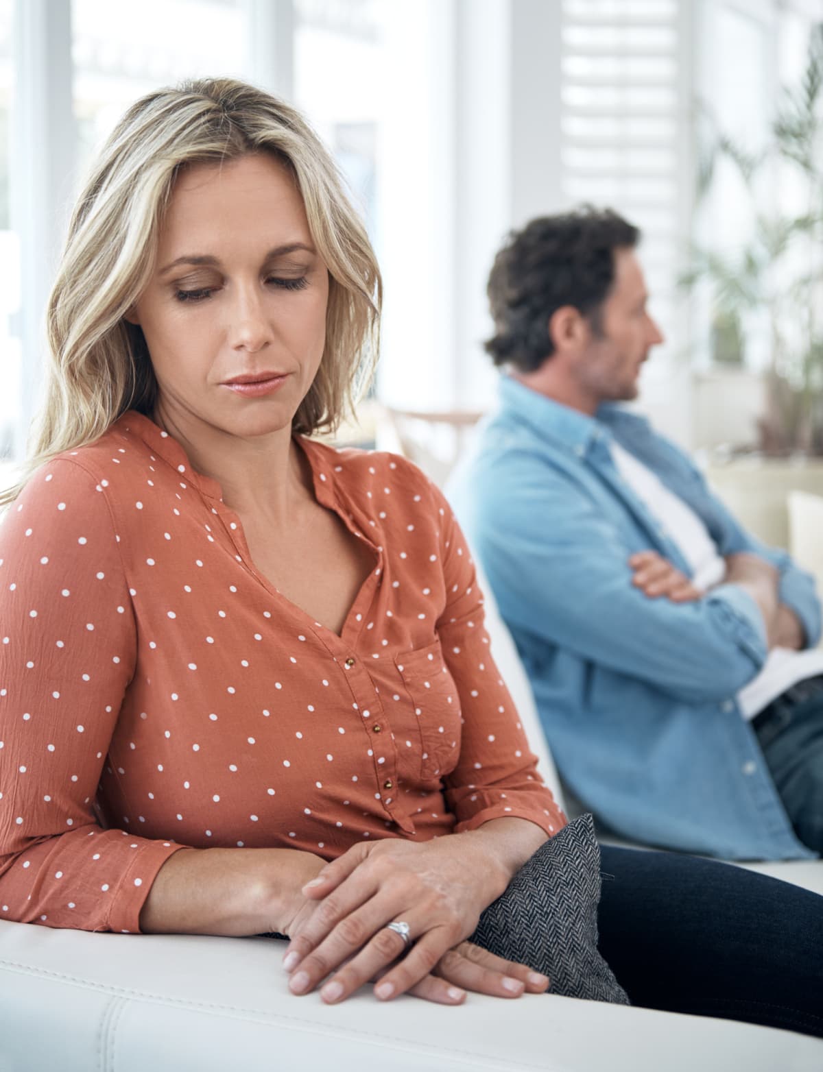 A woman with a sad expression on her face sits on a couch next to her partner who has his arms crossed