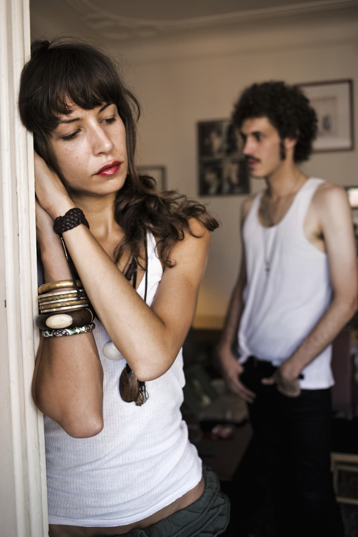 A woman leans on a doorframe looking downcast while a man in the background is looking somewhere else