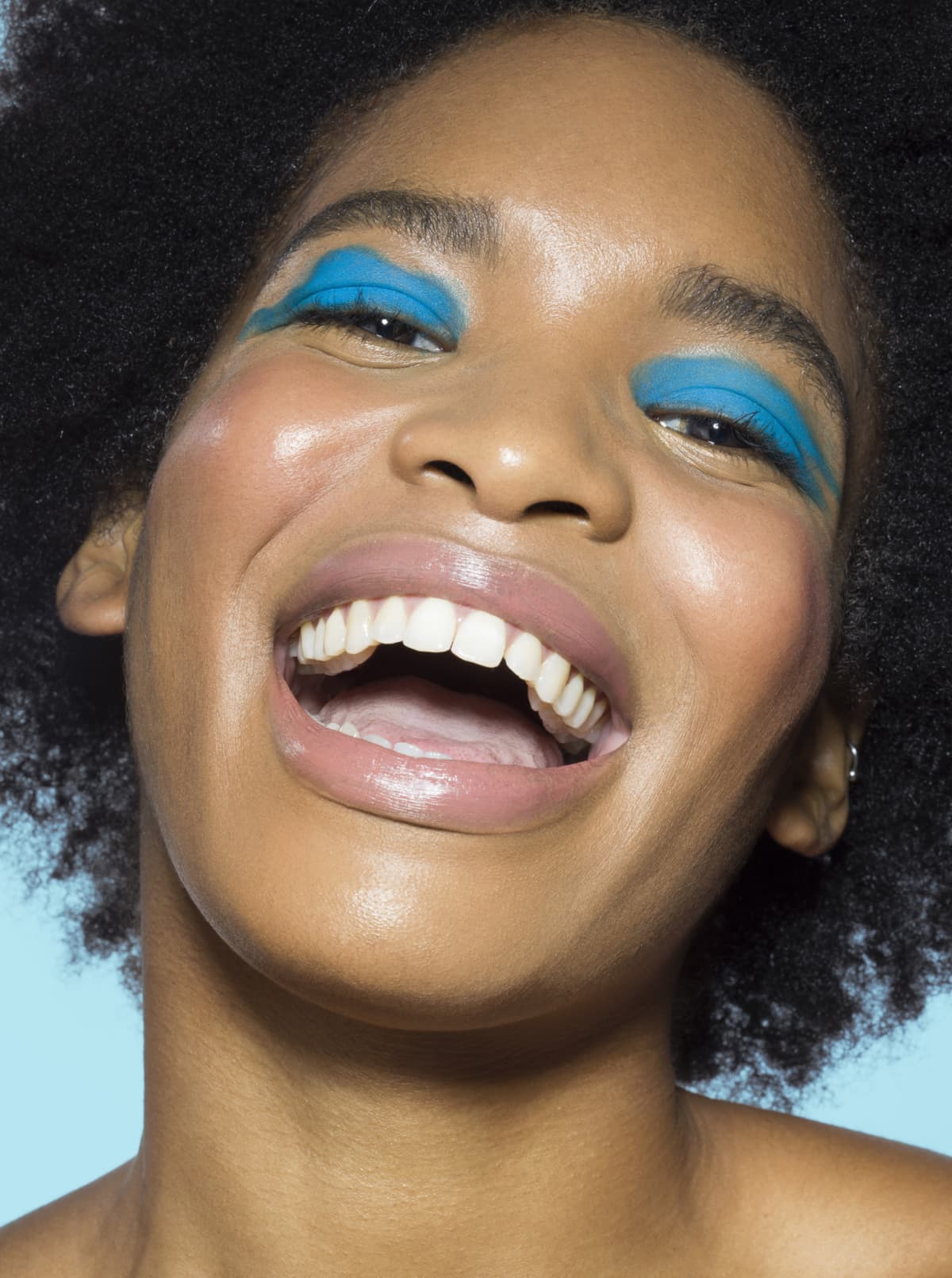 Close-up of young mixed-race woman looking to camera, laughing, wearing blue eye make-up with turquoise background