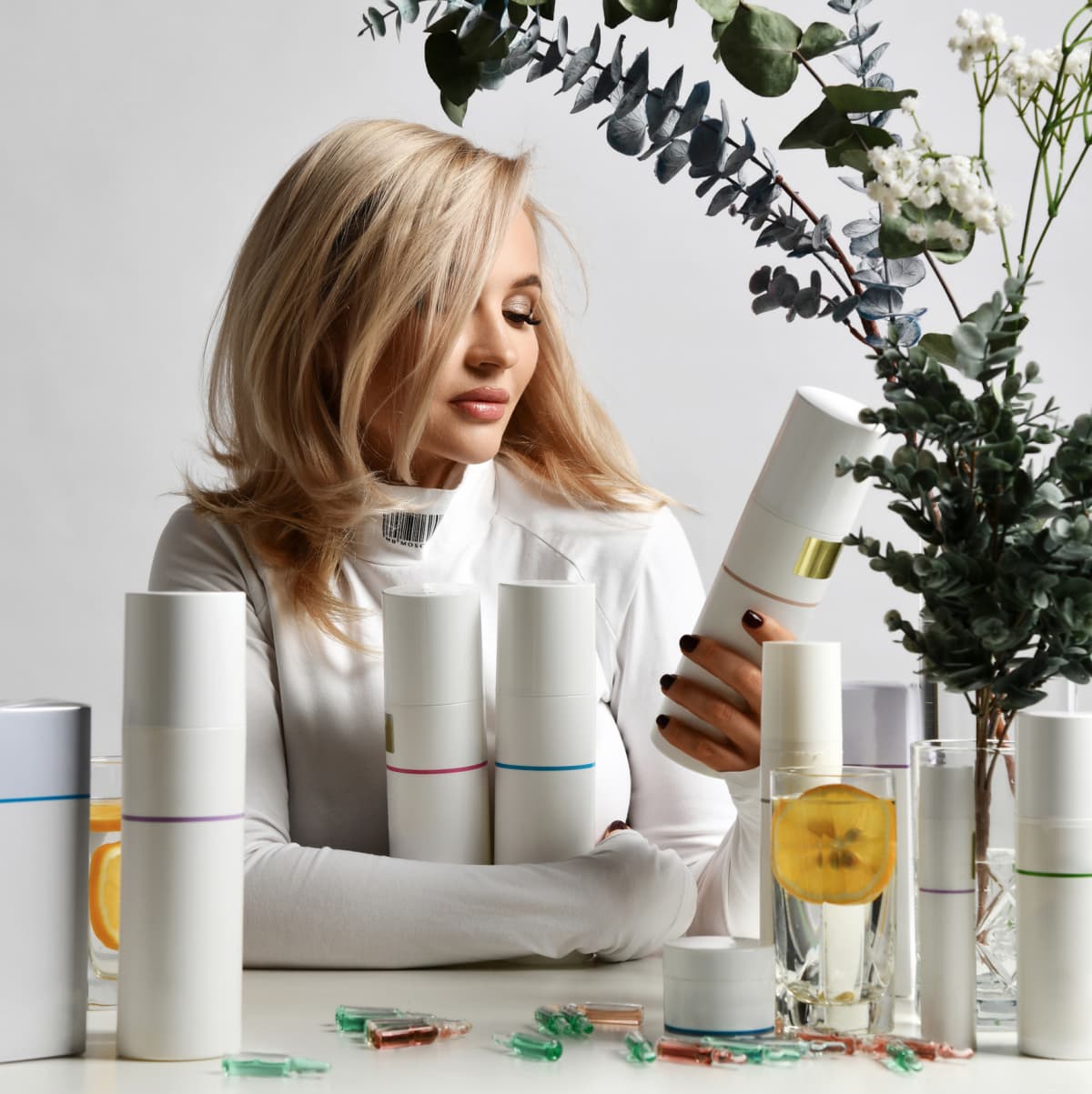 Rich blonde woman in white stylish pullover sits at table with set of jars and tubes of cosmetics, holding one in hand and looking at it. Beautiful woman, hair and skincare style concept, fashion