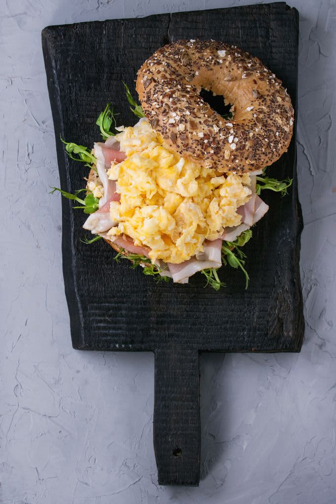 Opened Whole Grain bagels with scrambled eggs, pea sprout and prosciutto ham on wood plate over wooden textured background. (Photo by: Natasha Breen/REDA&CO/Universal Images Group via Getty Images)