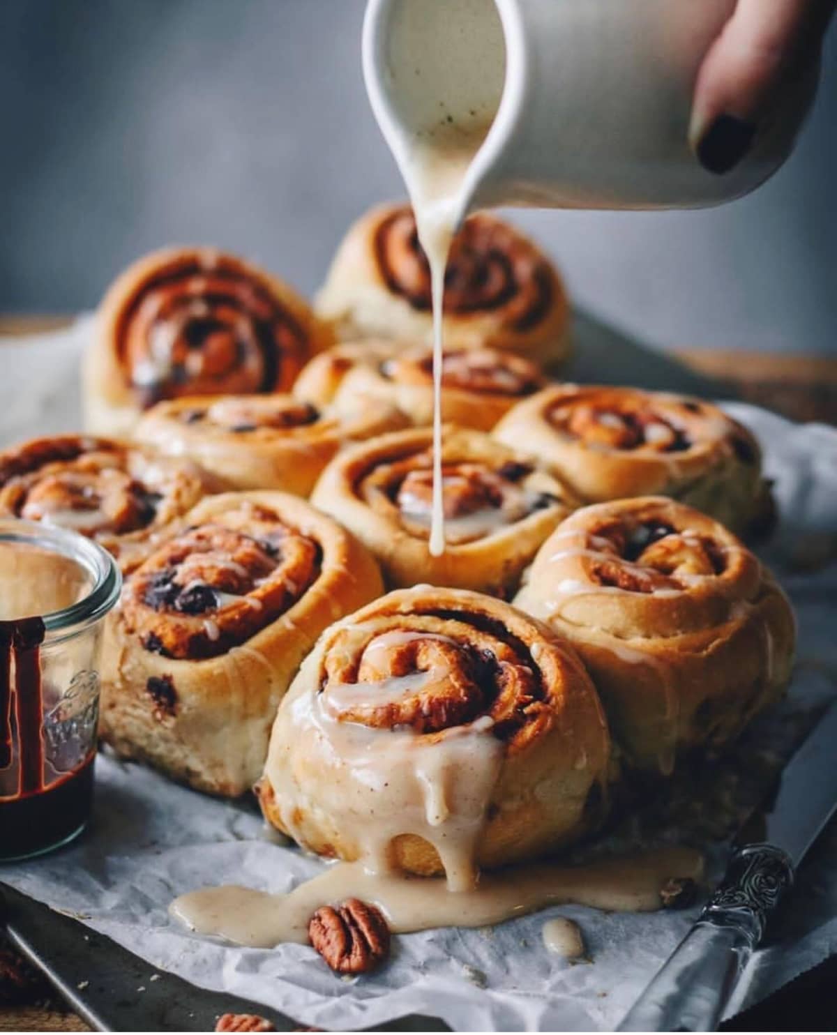 Icing being poured over a sheet tray of cinnamon buns.