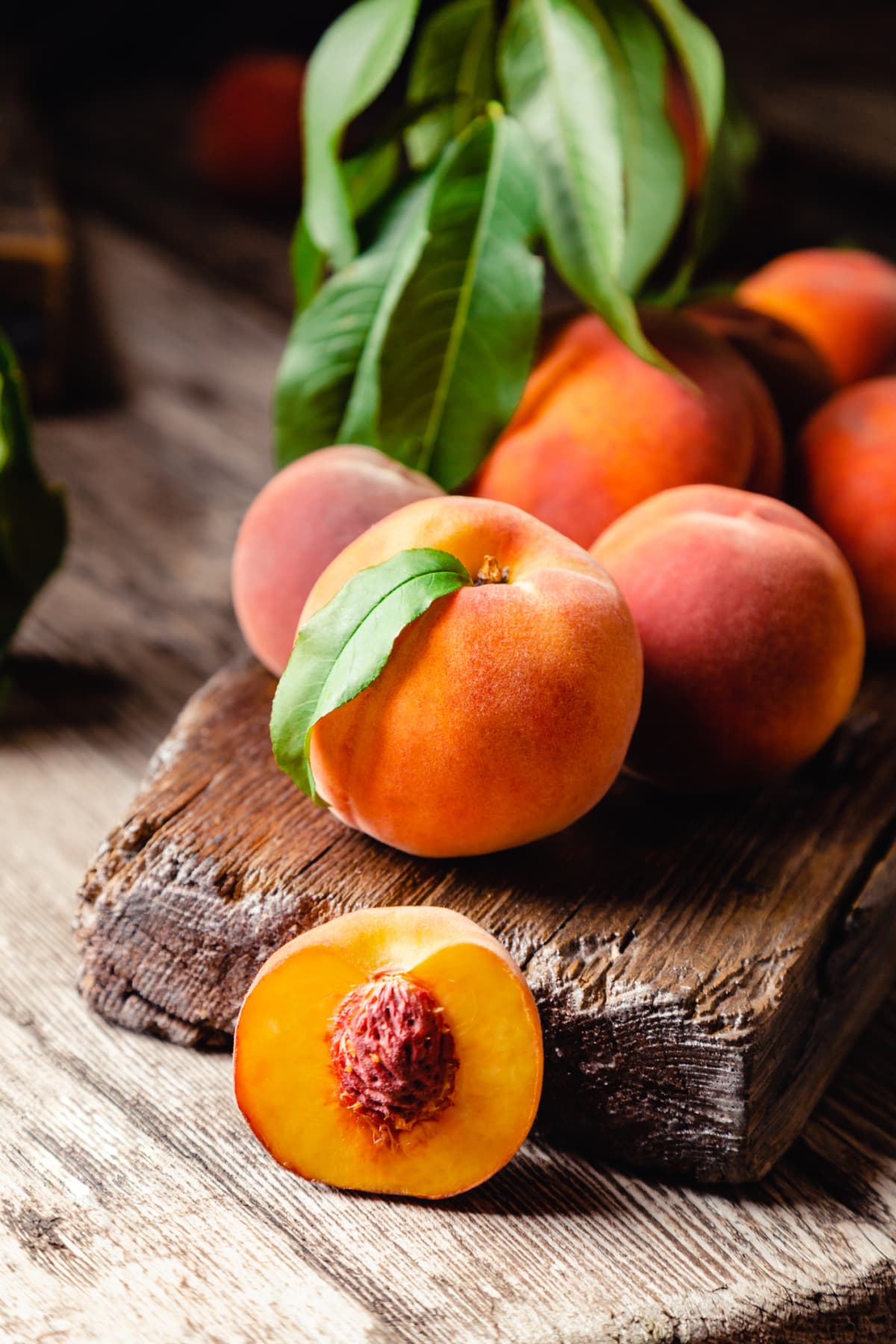 Peaches with leaves on dark wooden board with peach in halves with peach seed stone.