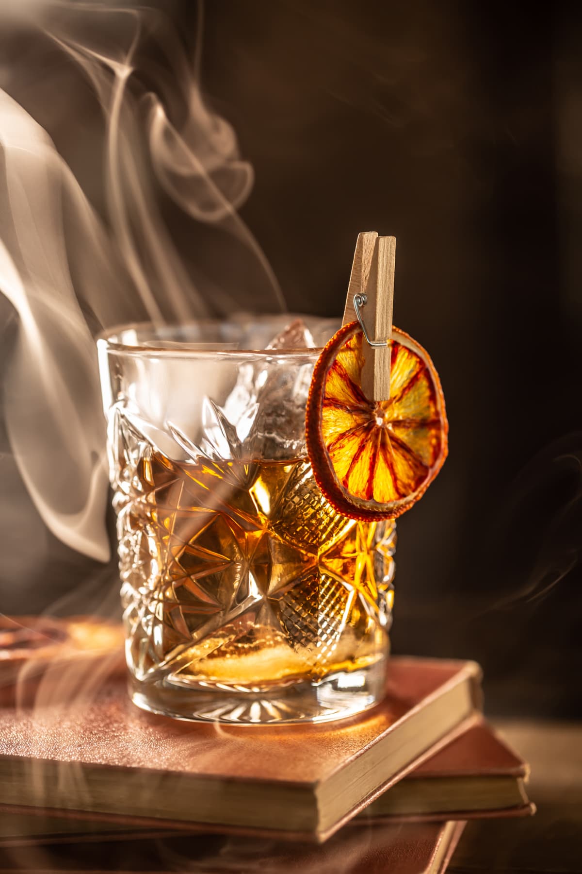 Smoked old fashioned rum cocktail with cubes of ice around on a dark background.