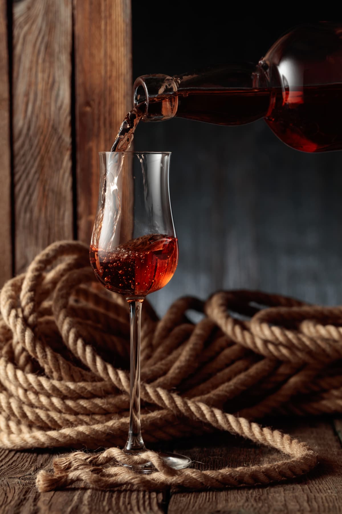 Dark alcohol pouring from bottle into a snifter. Old wooden background with hemp rope.