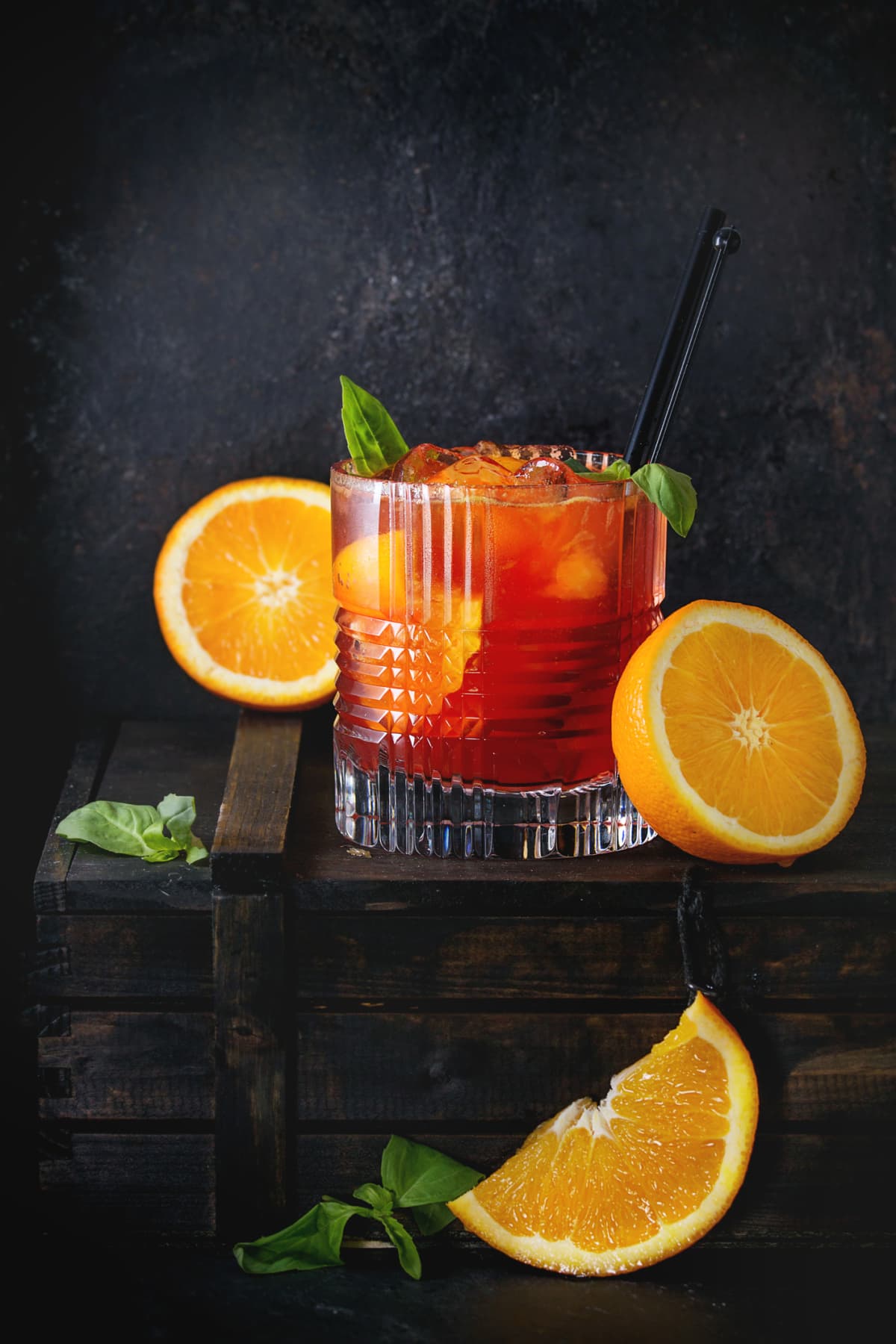 Alcohol cocktail Negroni with sliced orange and basil leaves, served on wooden box over black background.