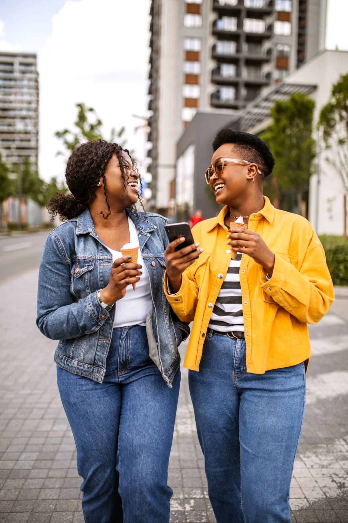 Two young women in a city walk on a sunny summer day, eating ice cream, using smart phone while walking, for social media and text messaging.