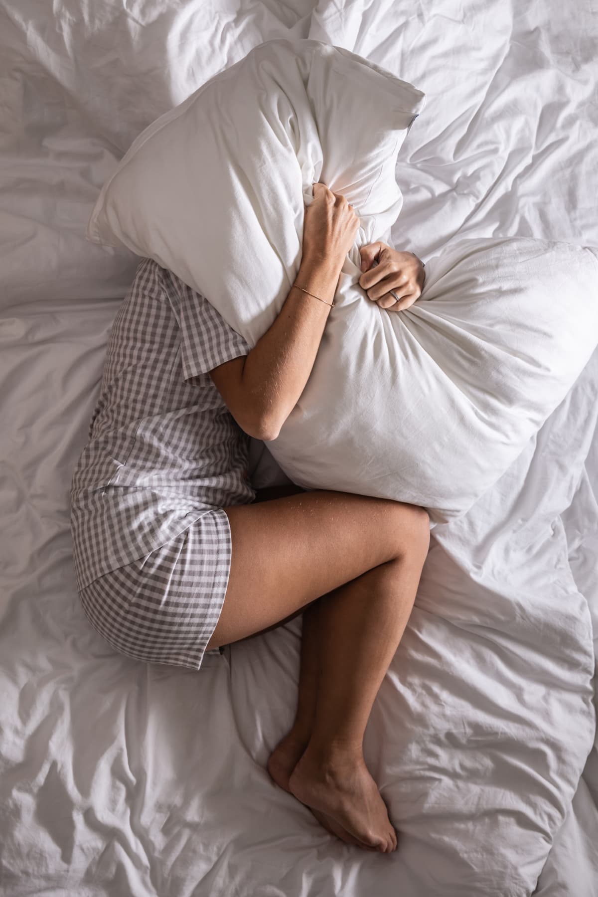 Woman in pyjamas holds pillow over her head, trying to cover from light to get more sleep.