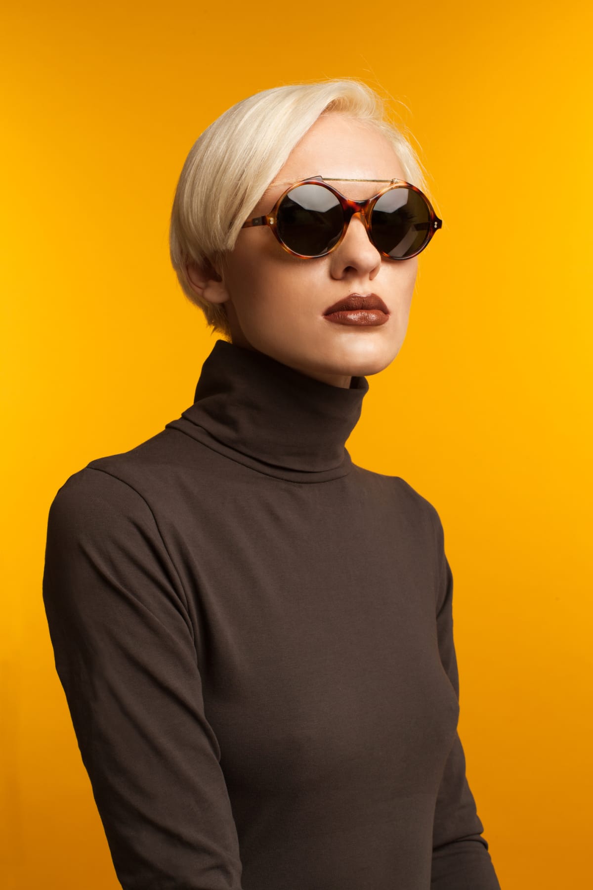 Portrait of blond girl wearing sunglasses and brown sweater. Shot in studio on yellow background.