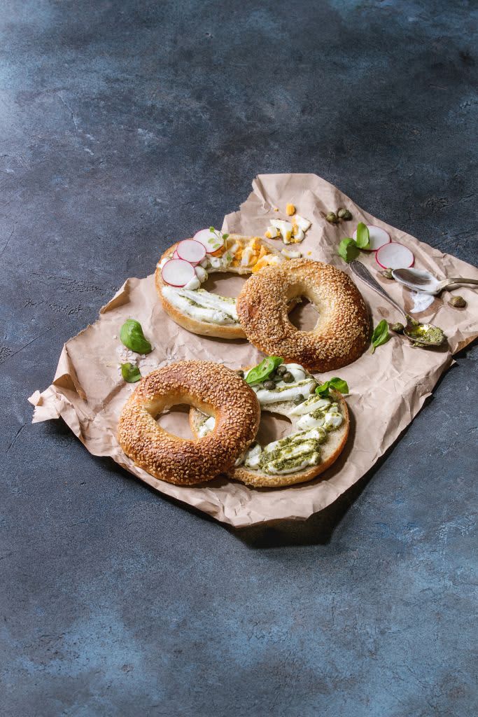 Variety of homemade bagels with sesame seeds, cream cheese, pesto sauce, eggs, radish, herbs served on red crumpled paper with ingredients above over grey texture background Top view, space. (Photo by: Natasha Breen/REDA&CO/Universal Images Group via Getty Images)