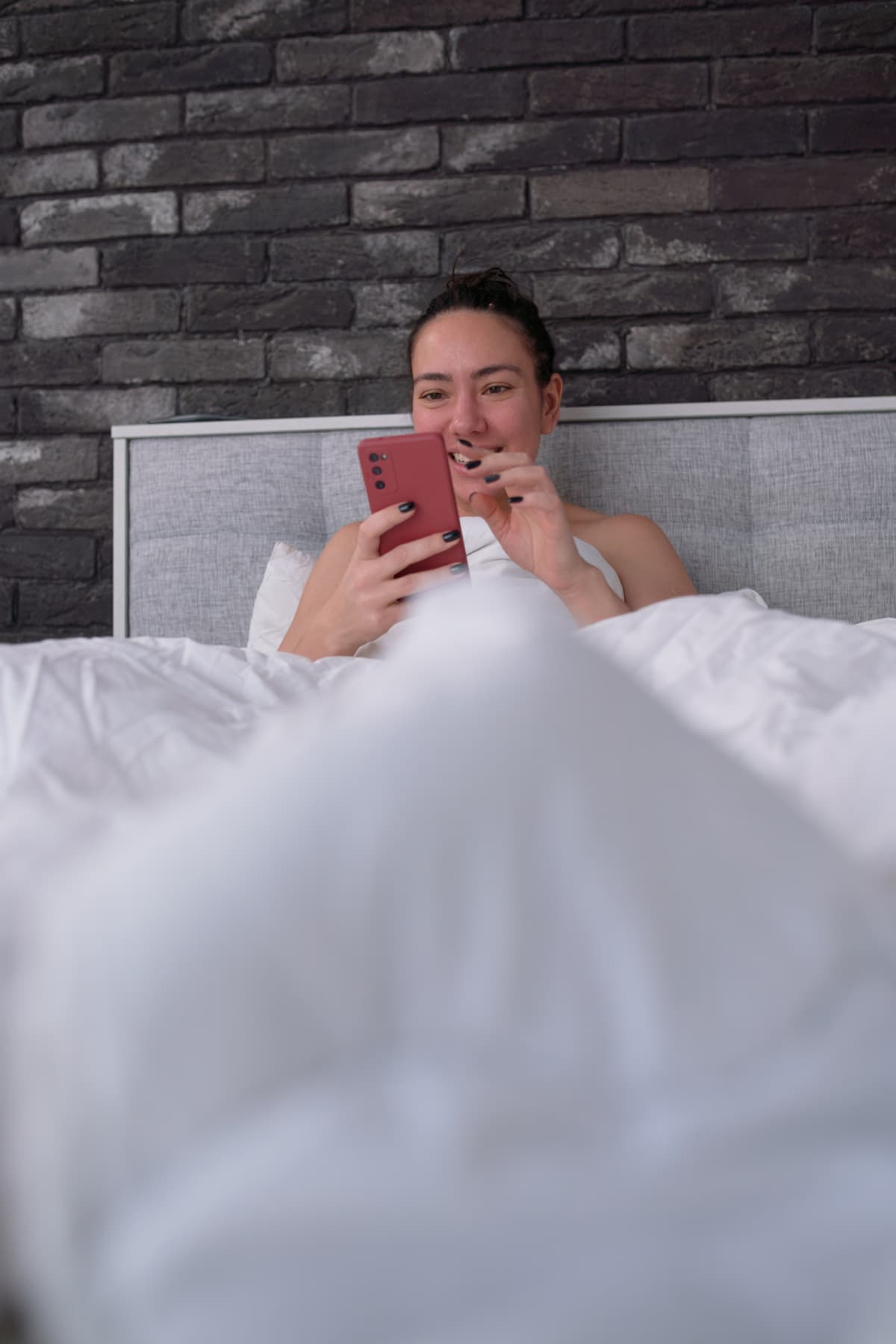 Happy female smiling and browsing social media on cellphone while resting in bed near brick wall in morning at home.