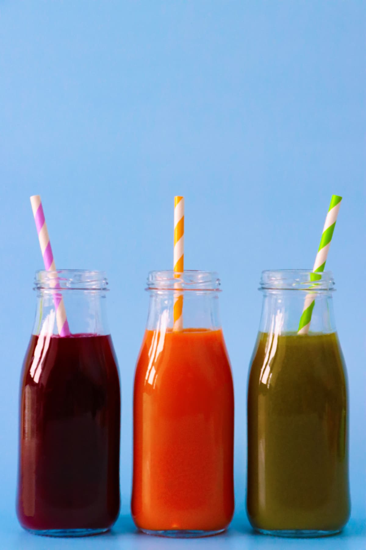Three different fruit vegetable smoothies served in vintage bottles with striped straws featuring a blue background