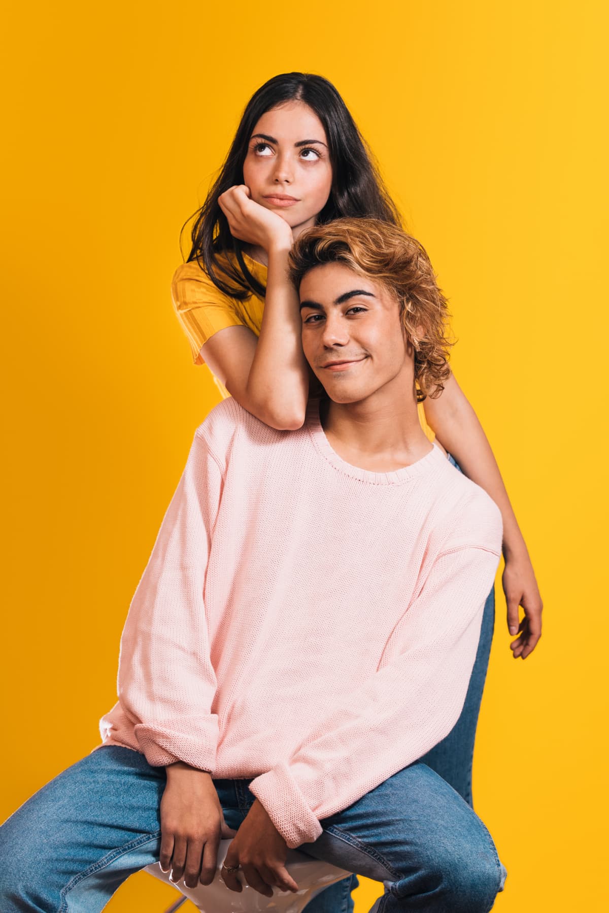 Young couple posing against a bright yellow background, the woman is standing behind the seated man rolling her eyes.