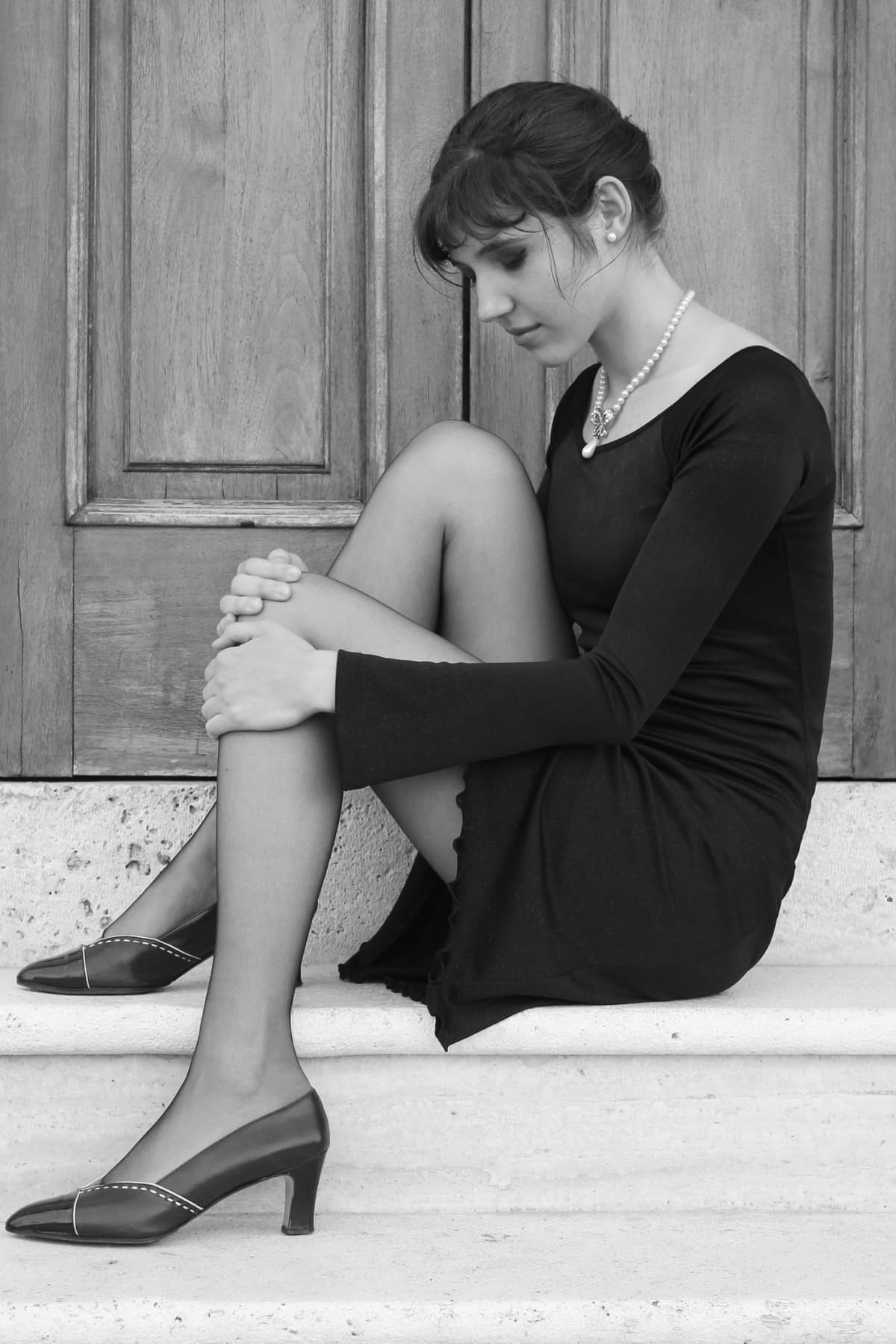 Black and white photo of woman in black dress and pantyhose sitting in front of door