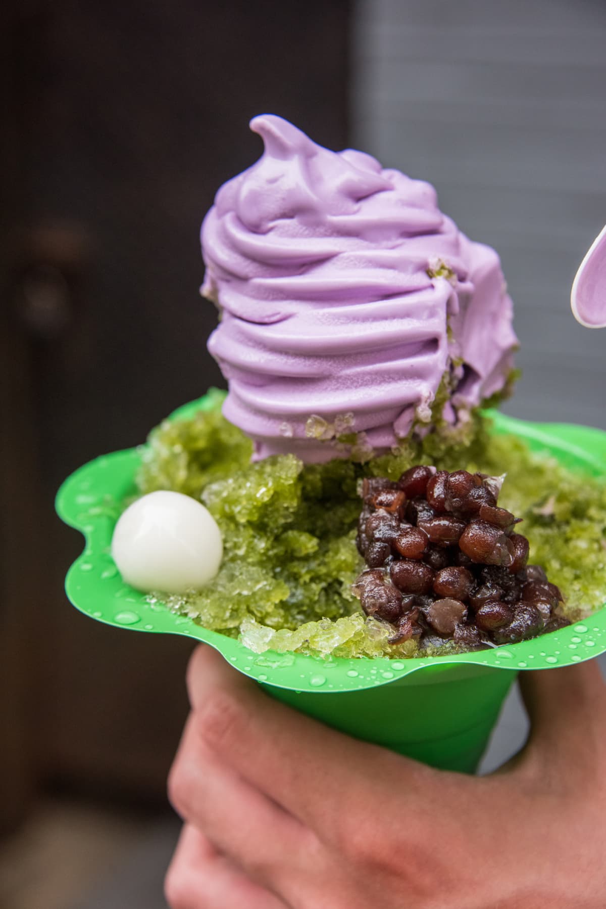 Close up of a hand holding a take out cone of matcha shaved ice topped with soft serve ube ice cream, red bean and mochi