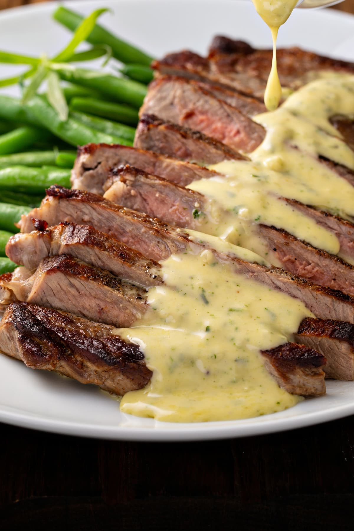 Sliced steak on a white plate with bearnaise sauce made with tarragon with vegetables in the background