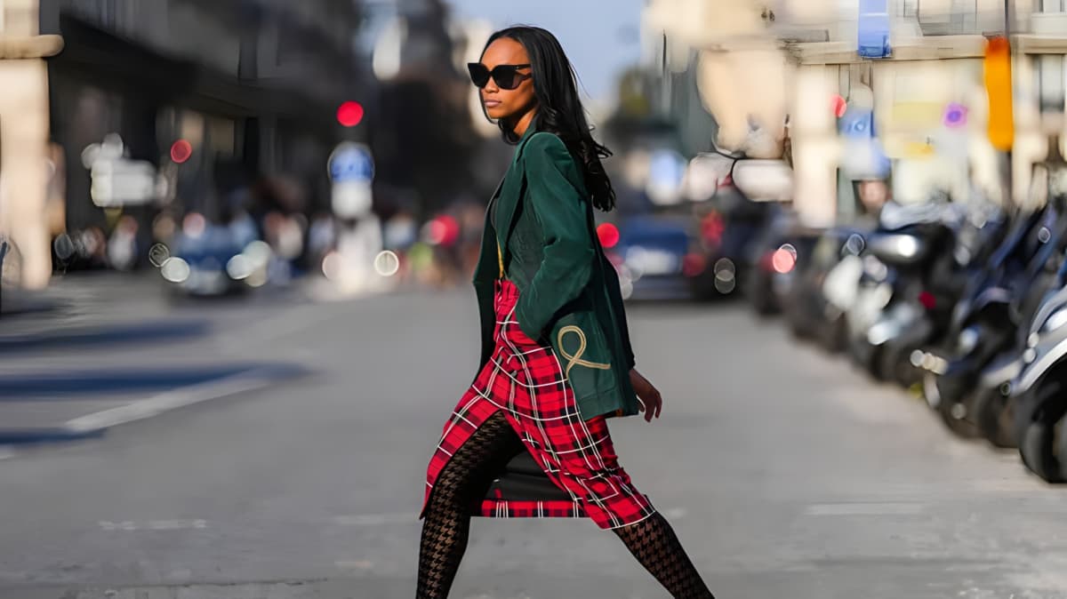 A woman in a green coat and tartan skirt walking on the road. 